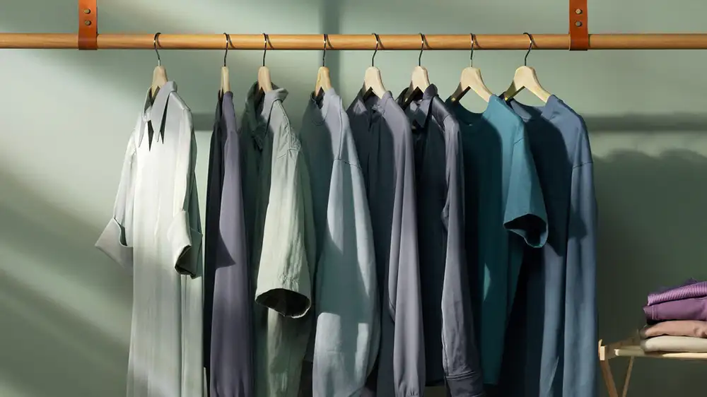 Na drewnianym wieszaku wiszą różne koszule i t-shirty w odcieniach szarości i niebieskiego. Obok na półce leżą złożone ubrania.