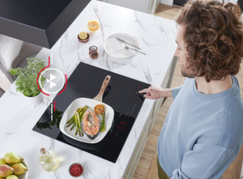 Osoba stoi przy płycie indukcyjnej, na której znajduje się patelnia z jedzeniem. Obok płyty widoczne są różne produkty spożywcze i naczynia na blacie kuchennym.
