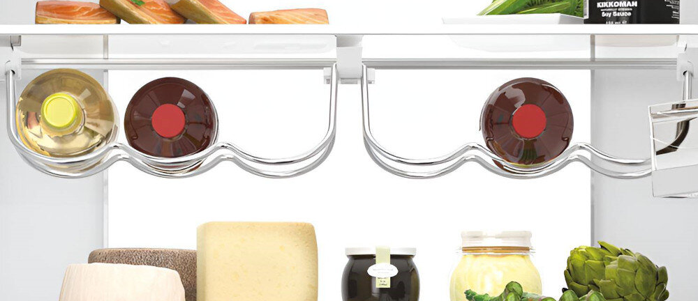 Wnętrze lodówki z widocznymi półkami na butelki, na których znajdują się dwie butelki wina. Na dolnej półce umieszczone są różne produkty spożywcze, w tym słoiki i warzywa.