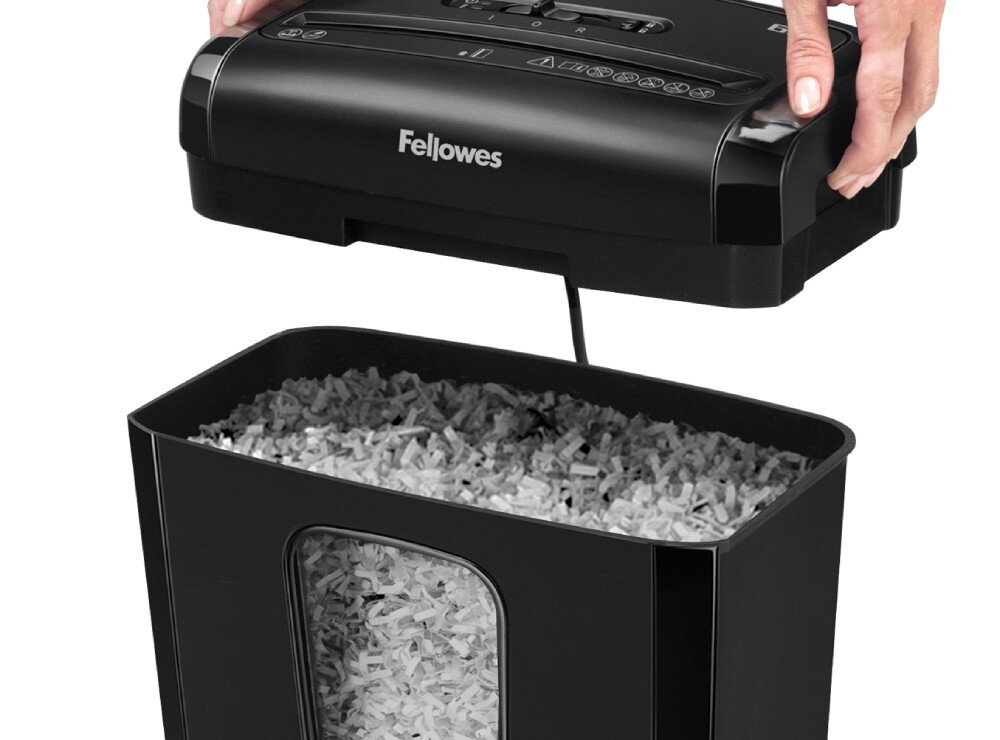 Czarna niszczarka z logo \'Fellowes\' na górnej części, z otwartym pojemnikiem wypełnionym pociętym papierem. Dwie ręce trzymają górną część urządzenia.