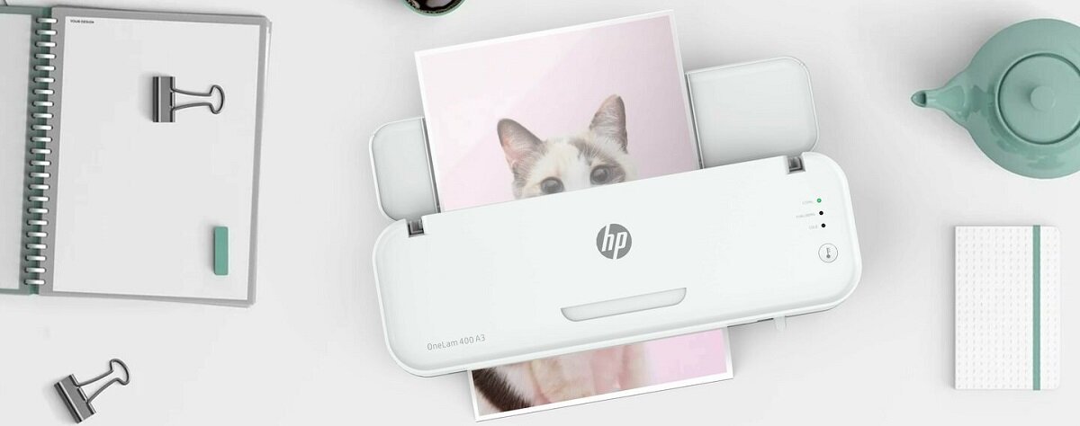 Na obrazie znajduje się laminator z logo HP, przez który przechodzi zdjęcie kota. Obok widoczne są notatnik, spinacze i dzbanek.