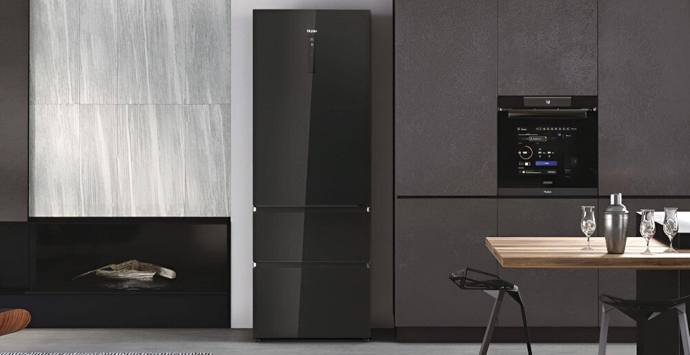 Czarna lodówka z trzema drzwiami stoi w nowoczesnej kuchni obok zabudowanego piekarnika. Na lodówce widoczne jest logo \'Haier\'.