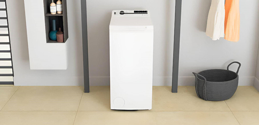 Na obrazie znajduje się biała pralka ładowana od góry, ustawiona w jasnym pomieszczeniu. Obok pralki widoczny jest kosz na pranie oraz półka z kosmetykami.