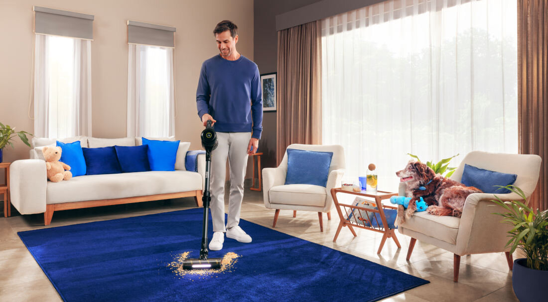 Osoba używa odkurzacza na niebieskim dywanie w salonie z jasnymi meblami i dużym oknem. Na kanapie obok siedzi pies, a na stoliku znajduje się szklanka z napojem.