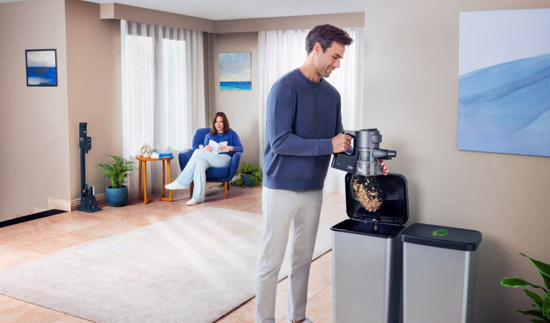 Mężczyzna opróżnia pojemnik odkurzacza do kosza na śmieci w jasnym, nowoczesnym wnętrzu. W tle widać osobę siedzącą na fotelu z książką.