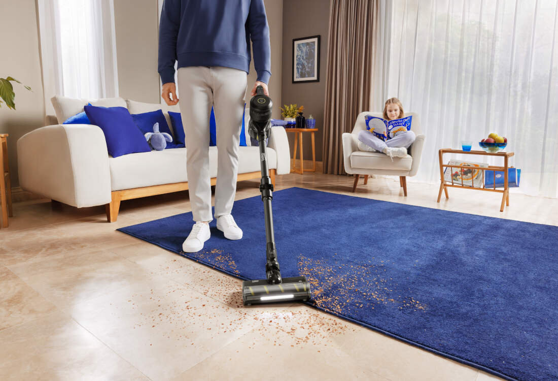 Osoba używa odkurzacza pionowego na niebieskim dywanie, na którym znajdują się rozsypane drobinki. W tle widać dziecko siedzące na fotelu z książką.
