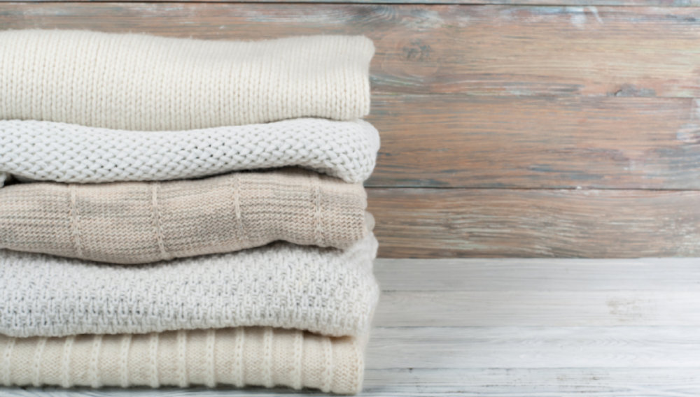 Stos złożonych swetrów w odcieniach beżu i bieli na drewnianym tle.