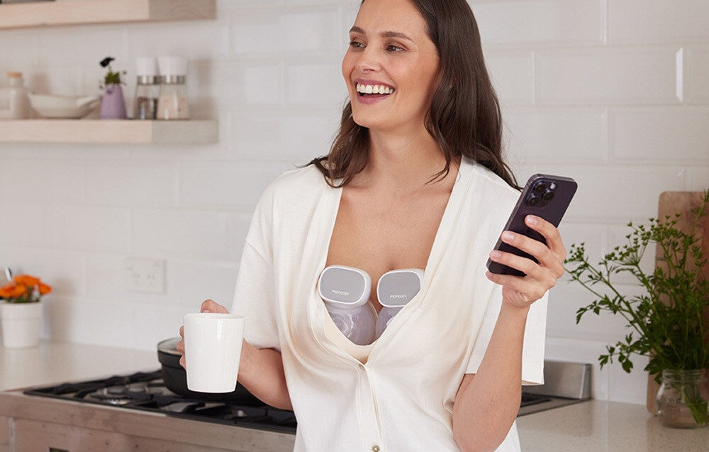 Osoba trzymająca kubek i telefon, z widocznym laktatorem MOMCOZY umieszczonym pod ubraniem. W tle kuchnia z białymi kafelkami i rośliną.