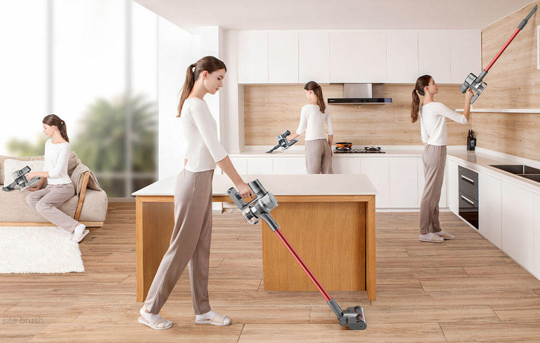 Na obrazie widać kilka osób używających odkurzacza bezprzewodowego w nowoczesnej kuchni. Odkurzacz ma szarą obudowę i czerwoną rurę ssącą.