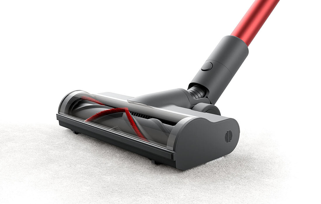 Na obrazie widoczna jest głowica odkurzacza z przezroczystą obudową, przez którą widać szczotkę z czerwonymi elementami. Odkurzacz znajduje się na jasnym dywanie.