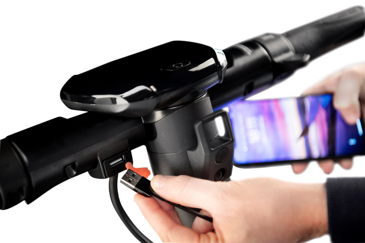 Osoba trzyma telefon komórkowy i podłącza kabel USB do portu na kierownicy hulajnogi elektrycznej. Kierownica jest czarna z widocznym wyświetlaczem i przyciskami.