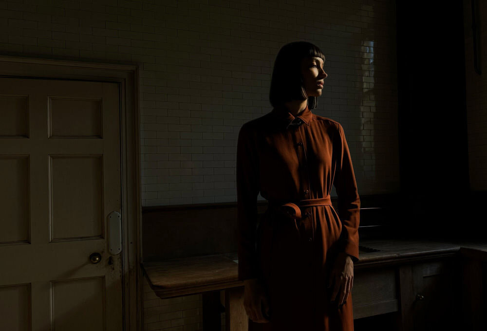 Osoba stoi w ciemnym pomieszczeniu przy drewnianym stole. Ściana jest wyłożona jasnymi płytkami, a obok znajduje się drzwi z klamką.