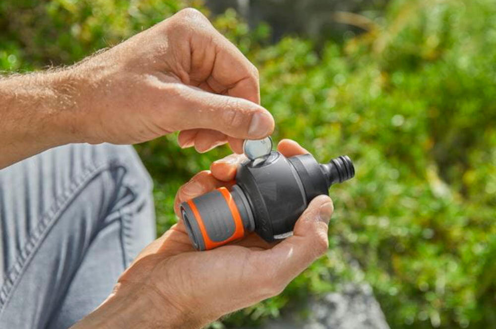 Osoba trzyma w rękach czarny licznik wody z pomarańczowym elementem, wkładając do niego baterię. W tle widoczna jest zielona trawa.