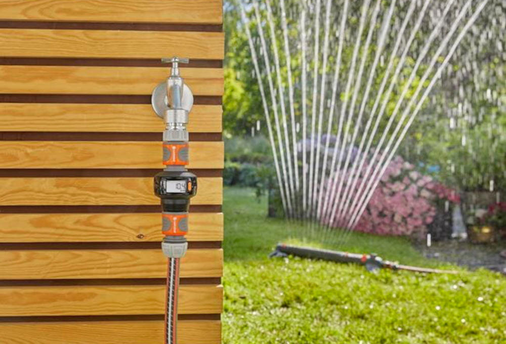 Na drewnianej ścianie zamontowany jest licznik wody z wężem ogrodowym, a w tle widać działający zraszacz na trawniku. Licznik ma wyświetlacz cyfrowy i elementy w kolorze pomarańczowym.
