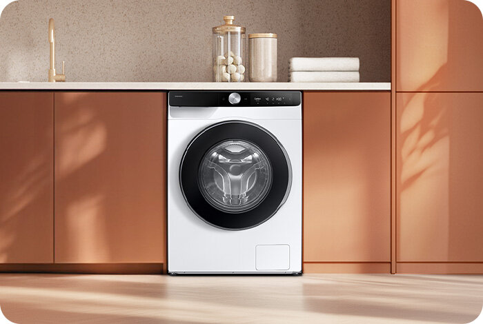 Pralka w zabudowie kuchennej z białym frontem i czarnym panelem sterowania. Obok znajdują się szafki oraz dekoracyjne słoiki i ręczniki.