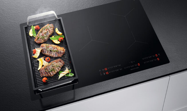 Czarna płyta indukcyjna z widocznymi panelami dotykowymi i grillowaną potrawą na patelni. Na płycie znajdują się cyfry i symbole kontrolne w kolorze czerwonym i białym.