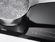 Na obrazie widoczna jest płyta indukcyjna z garnkiem pełnym gotującej się wody. Obok znajdują się dotykowe elementy sterujące z czerwonymi wskaźnikami cyfrowymi i symbolami.