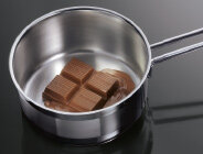 Stalowy rondel z kawałkami czekolady, które zaczynają się topić, umieszczony na ciemnej powierzchni.