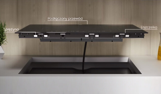 Obraz przedstawia płytę indukcyjną zamontowaną nad blatem z widocznymi elementami: sprężyną, podłączonym przewodem i uszczelką. Tło stanowi drewniana ściana.