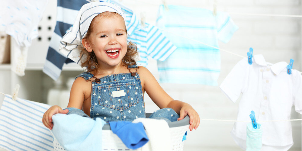 Dziecko siedzi w koszu na pranie, ubrane w ogrodniczki i chustkę na głowie. W tle widoczne są rozwieszone ubrania w jasnych kolorach.