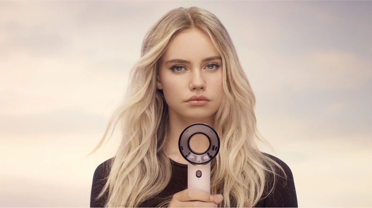 Modelka o długich blond włosach trzyma w dłoni przed sobą niedużą jasnoróżową suszarkę Dyson Supersonic Travel