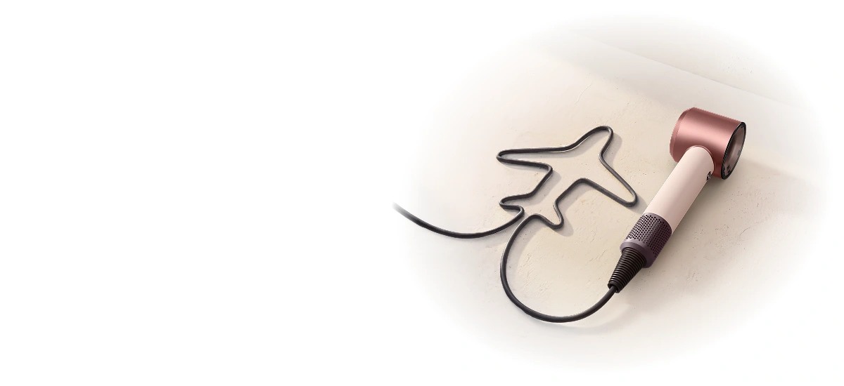 Jasnoróżowa suszarka Dyson Supersonic Travel leży na jasnym blacie. Jej kabel jest ułożony obok w kształcie samolotu