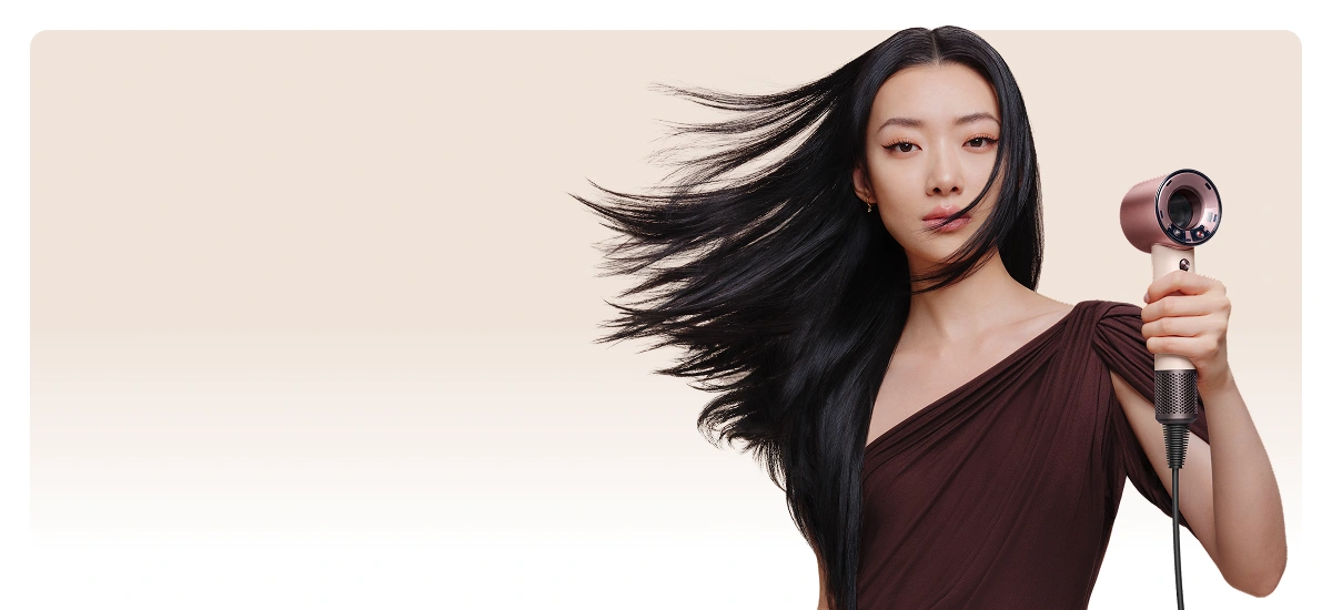 Modelka o długich, czarnych włosach trzyma w ręku przed sobą suszarkę i suszy włosy. Powietrze z suszarki rozwiewa jej włosy