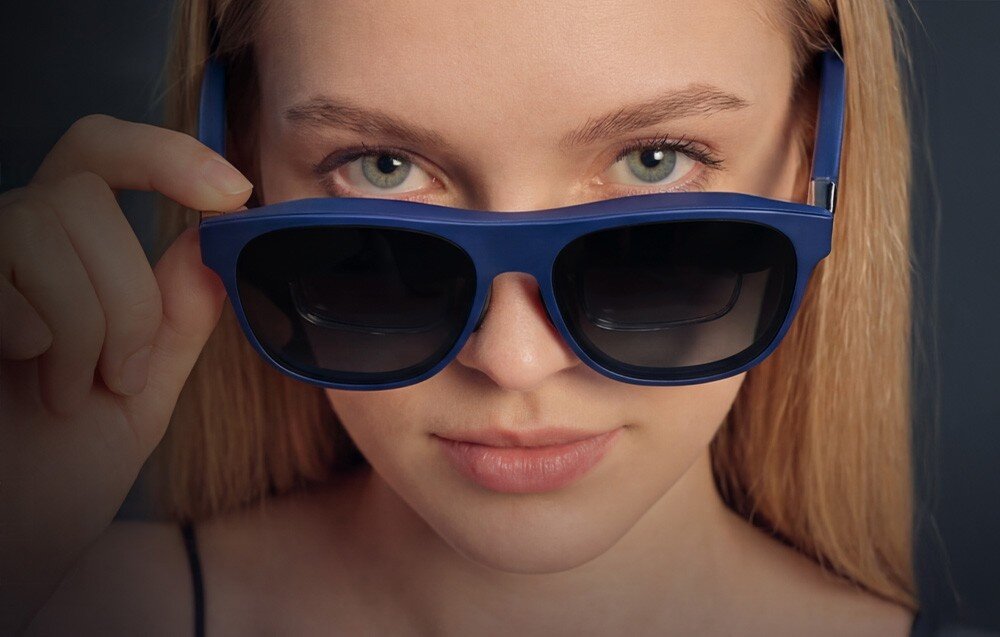 Okulary XREAL 1S Portret blondynki spoglądającej znad niebieskich oprawek okularów AR XREAL.