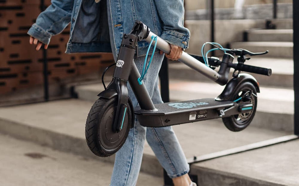 Osoba trzyma szarą hulajnogę elektryczną z widocznym logo \'MOTUS\'. Hulajnoga ma czarne koła i niebieskie akcenty na przewodach.