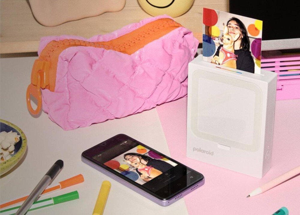 Drukarka POLAROID Hi-Print 3x3 Przenośna drukarka do zdjęć stojąca na biurku obok telefonu z wyświetlonym zdjęciem oraz przyborów szkolnych