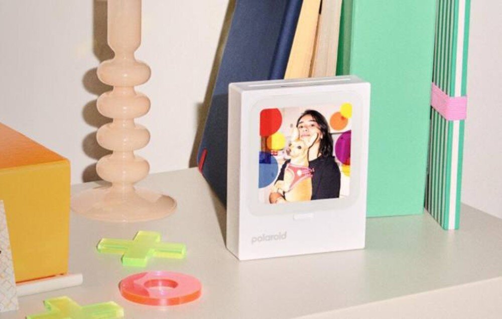 Drukarka POLAROID Hi-Print 3x3 Drukarka do zdjęć stojąca na półce obok książek oraz dekoracyjnych elementów