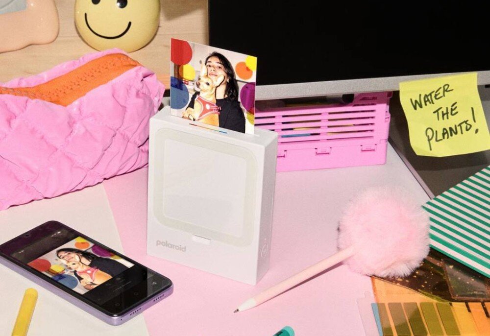 Drukarka POLAROID Hi-Print 3x3 Przenośna drukarka z wysuwającym się odbitką stojąca na biurku w otoczeniu telefonu i artykułów biurowych
