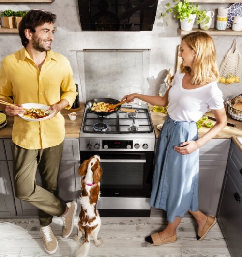 Dwie osoby gotują w kuchni, używając kuchenki gazowej z piekarnikiem. Obok nich stoi pies, a na blacie widoczne są różne akcesoria kuchenne i składniki.