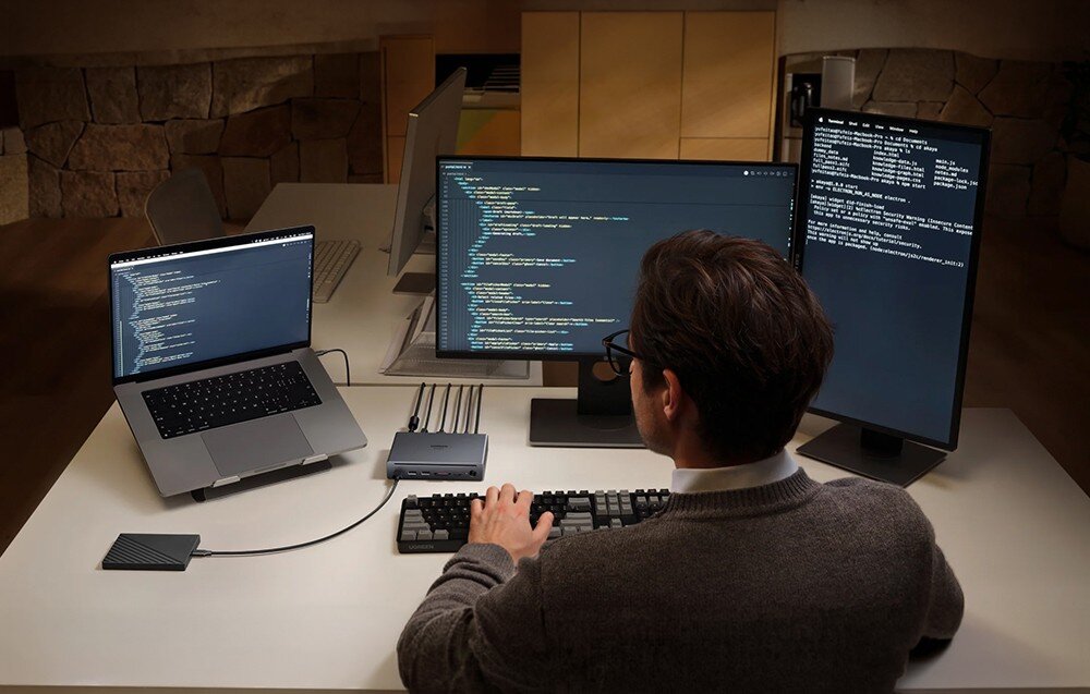 Osoba siedzi przy biurku z trzema monitorami i laptopem, na których wyświetlany jest kod programistyczny. Na biurku znajduje się stacja dokująca z kilkoma podłączonymi kablami.
