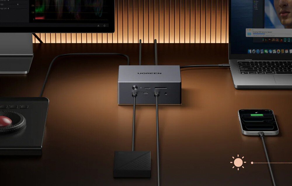 Na obrazie znajduje się stacja dokująca z logo UGREEN, podłączona do kilku urządzeń, w tym monitora i laptopa. Na biurku widoczny jest także smartfon z podłączonym kablem oraz urządzenie z dużym czerwonym przyciskiem.