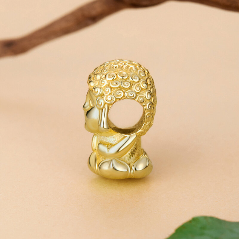 Złoty charms w kształcie Buddy z detalami na włosach, wykonany z metalu o błyszczącej powierzchni. Tło jest jednolite, w kolorze beżowym, z fragmentem gałązki i liścia.