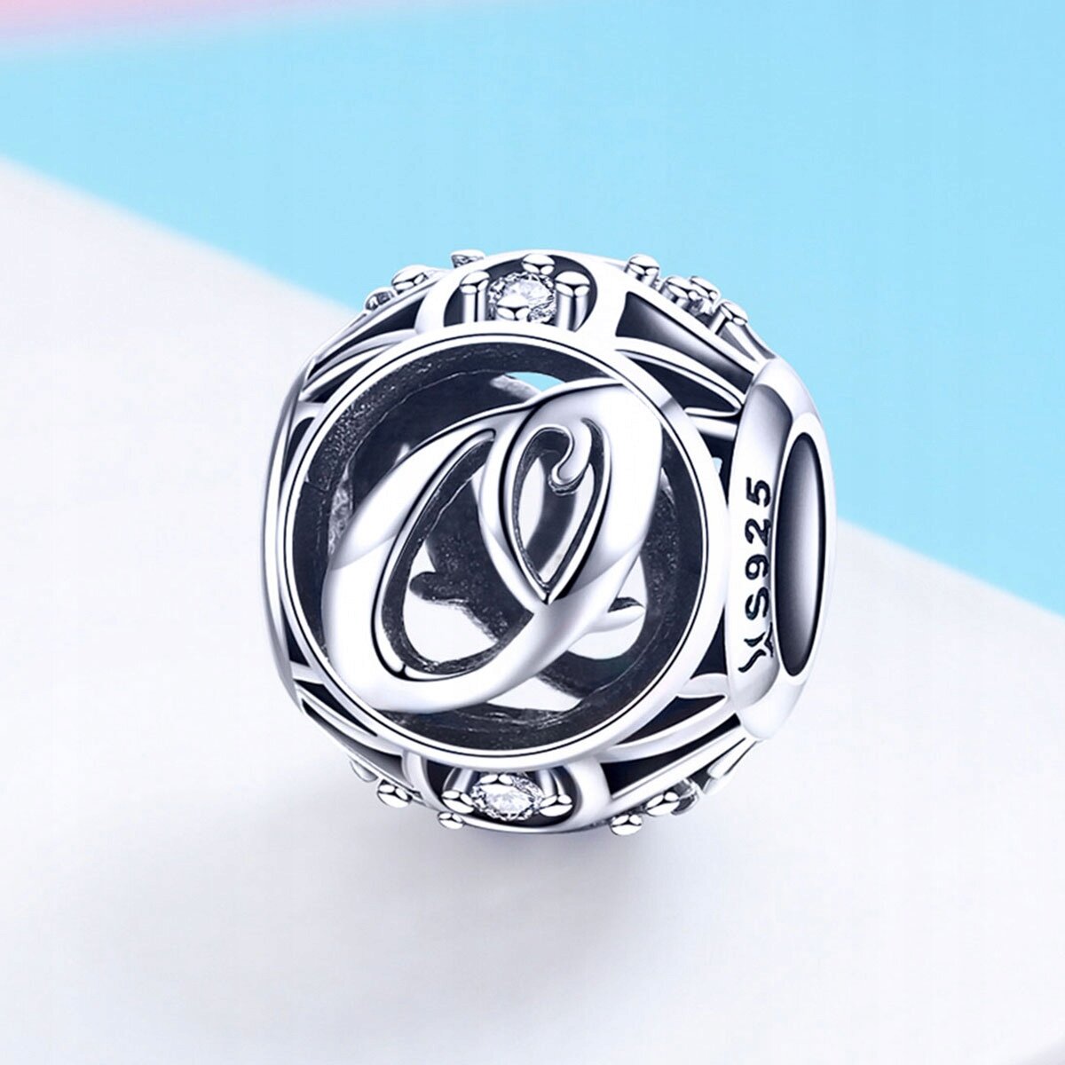 Srebrna zawieszka charms z literą \'O\', ozdobiona drobnymi kamieniami i oznaczeniem \'S925\'.