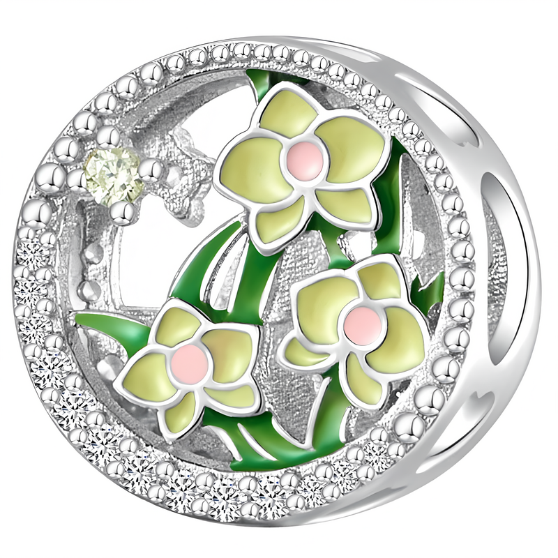 Srebrna zawieszka charms z motywem trzech kwiatów orchidei w kolorze żółtym z różowymi środkami, otoczona drobnymi, błyszczącymi elementami. W tle widoczne są zielone liście.