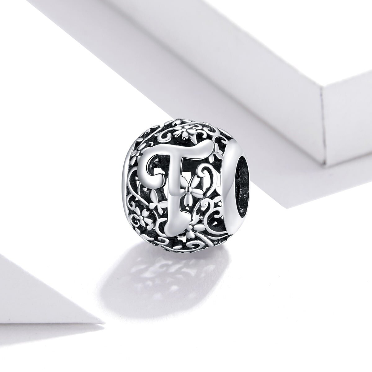 Srebrna zawieszka charms z literą \'T\' na ażurowym tle z kwiatowym wzorem. Produkt wykonany z metalu o wysokim połysku, prezentowany na jasnym tle.
