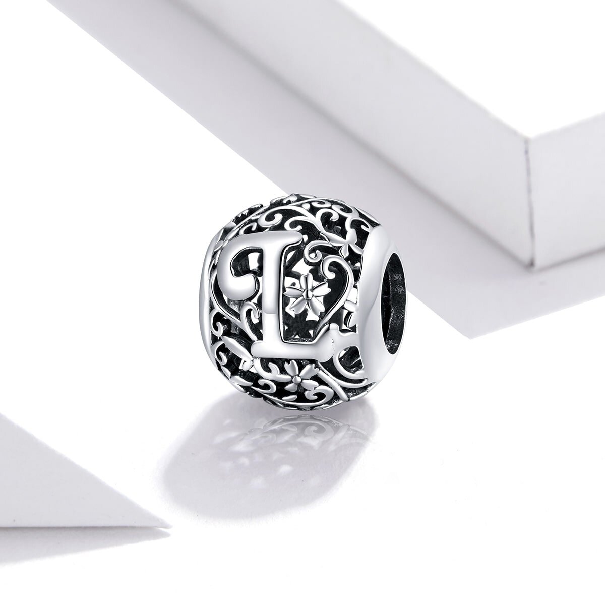 Srebrna zawieszka charms z literą \'L\' na tle ozdobnych wzorów roślinnych. Produkt jest umieszczony na białym tle.