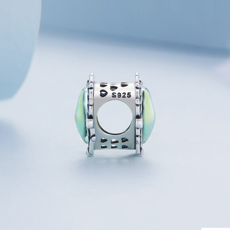 Srebrna zawieszka charms z oznaczeniem \'S925\' i ozdobnymi elementami w kolorze zielonym. Tło jest jasnoniebieskie.