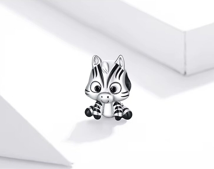 Srebrna zawieszka w kształcie małej zebry z czarnymi paskami, siedząca na białym tle. Zebra ma duże oczy i wyraźne uszy.