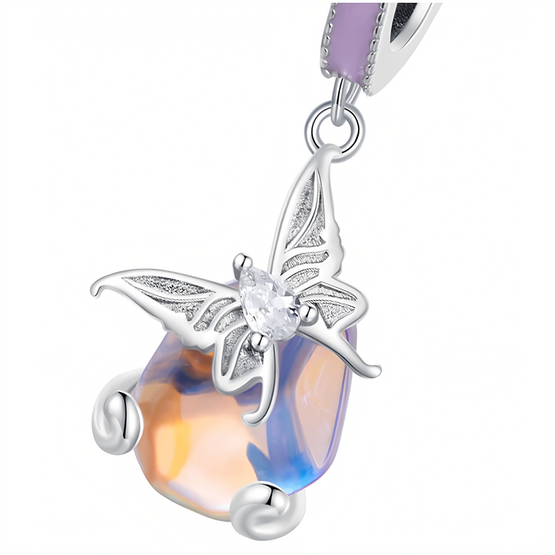 Zawieszka charms z motywem motyla, wykonana z opalizującego materiału w odcieniach fioletu i pomarańczu. Motyl ma srebrne skrzydła z detalami i centralny, przezroczysty kamień.