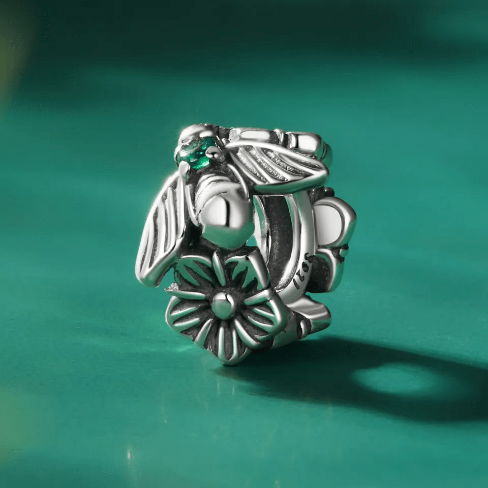 Srebrna zawieszka charms z motywem kwiatu i owada, ozdobiona zielonym kamieniem. Widoczny napis \'S925\'.