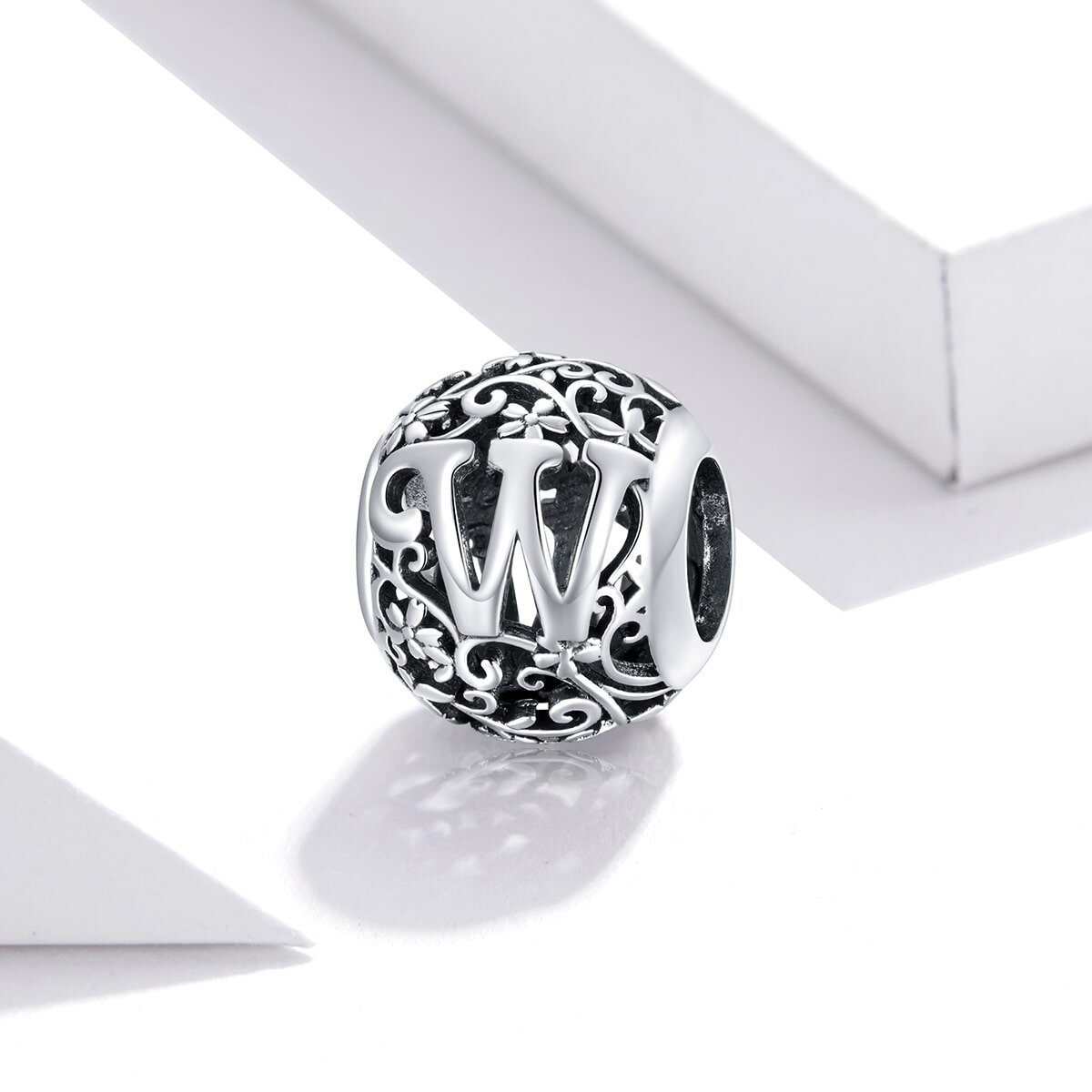 Srebrna zawieszka charms z literą \'W\' na ażurowym tle z motywem roślinnym. Produkt jest sfotografowany na białym tle.