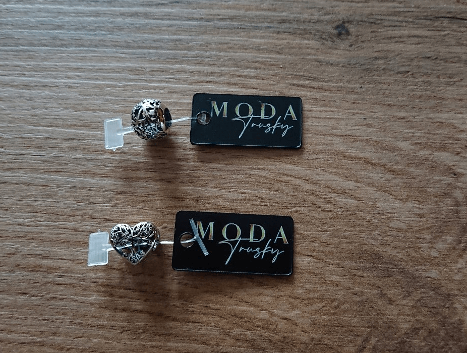 Na drewnianej powierzchni leżą dwie zawieszki charms w kształcie serca z ażurowym wzorem, każda z przyczepioną czarną etykietą z napisem \'MODA Trusky\'.