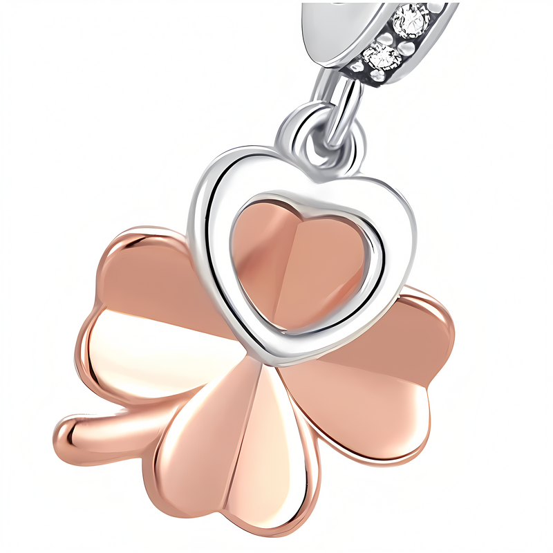 Zawieszka charms w kształcie koniczynki z sercem, wykonana z metalu w kolorze różowego złota i srebra. Górna część zawieszki ozdobiona jest małymi, błyszczącymi kamieniami.