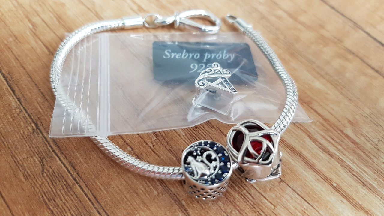 Na drewnianej powierzchni leży srebrna bransoletka z dwoma charmsami, z których jeden ma wzór czerwonej róży. Obok znajduje się przezroczysta torebka z napisem \'Srebro próby 925\'.