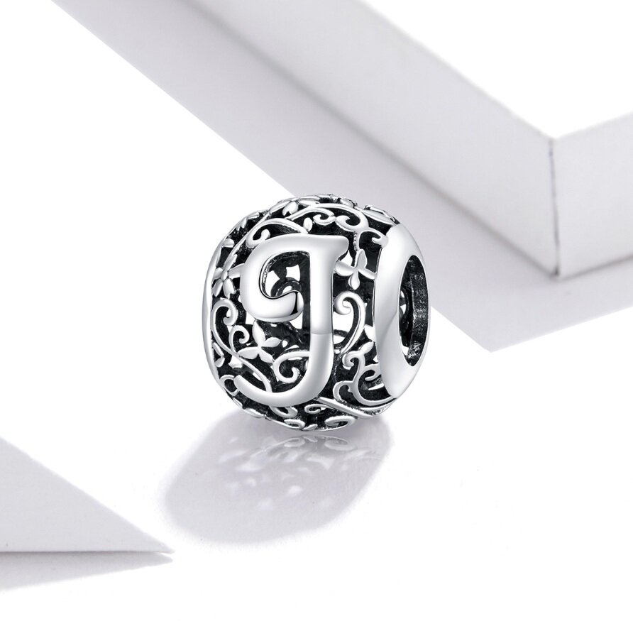 Srebrna zawieszka charms z literą \'J\' na ażurowym tle z motywem roślinnym. Produkt wykonany z metalu o błyszczącej powierzchni.