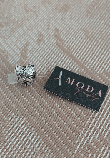 Na zdjęciu znajduje się srebrny koralik z wzorem serc oraz czarna karta z napisem \'MODA Trusky\'.
